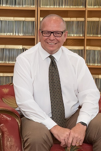 A man with glasses, sitting on a couch and smiling at the camera.