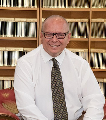 The image is a portrait of an older man with glasses, wearing a white shirt and tie, sitting in front of a shelf filled with books.