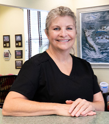 The image shows a woman standing behind a counter, smiling at the camera. She is wearing a black shirt and has short hair.