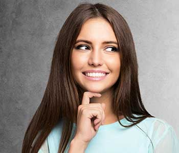 The image shows a smiling woman with her hand on her chin, looking directly at the camera. She has long brown hair and is wearing a light blue top.