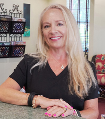 The image shows a woman with blonde hair wearing a black shirt and sitting behind a counter with various items on it.