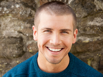 This is a color photograph of a smiling man with short hair, wearing a dark sweater and standing in front of a stone wall.