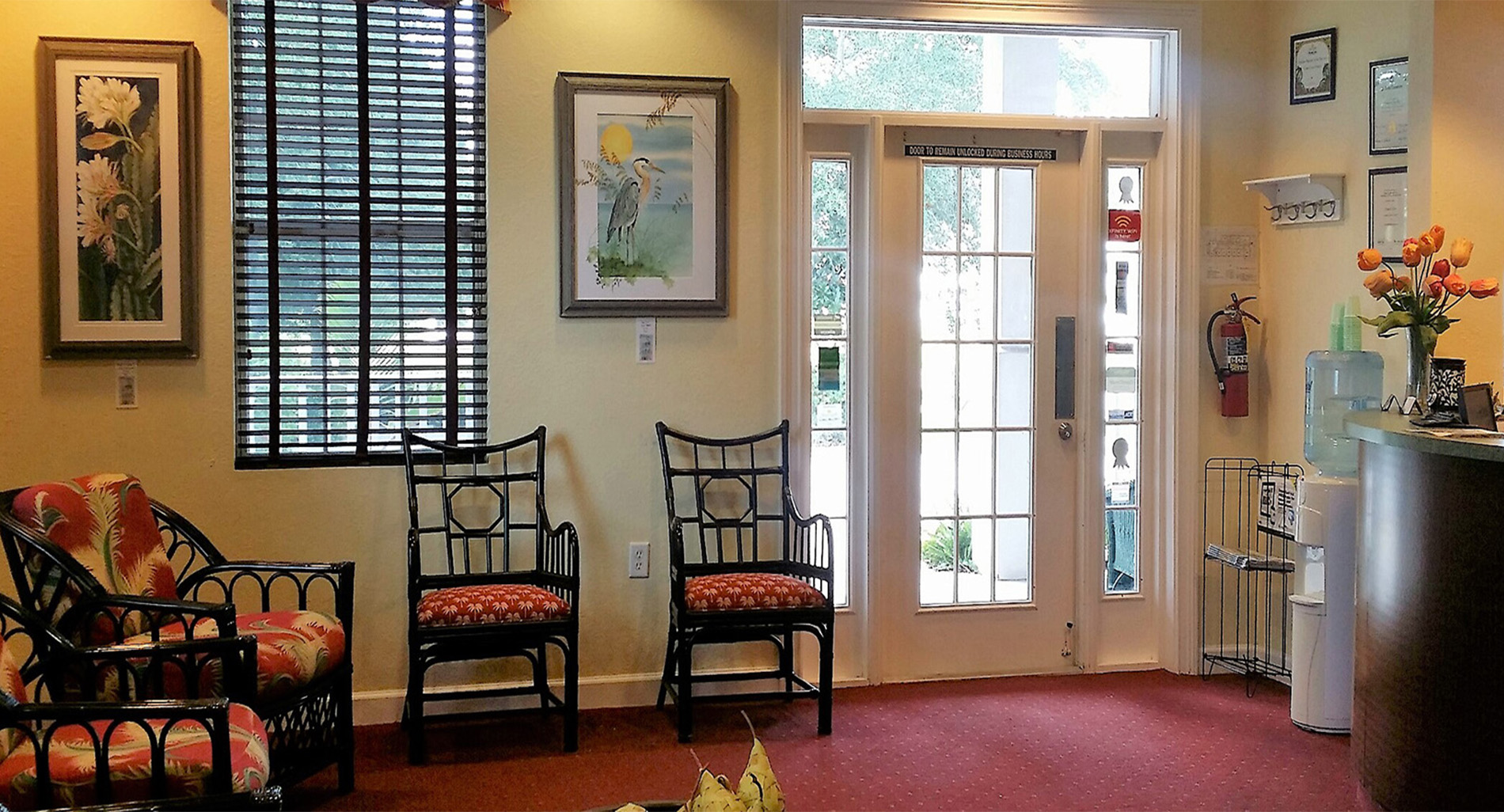 The image depicts an interior space that appears to be a waiting area or lobby, featuring a chair with a cushion, a painting on the wall, and a door with a glass panel.