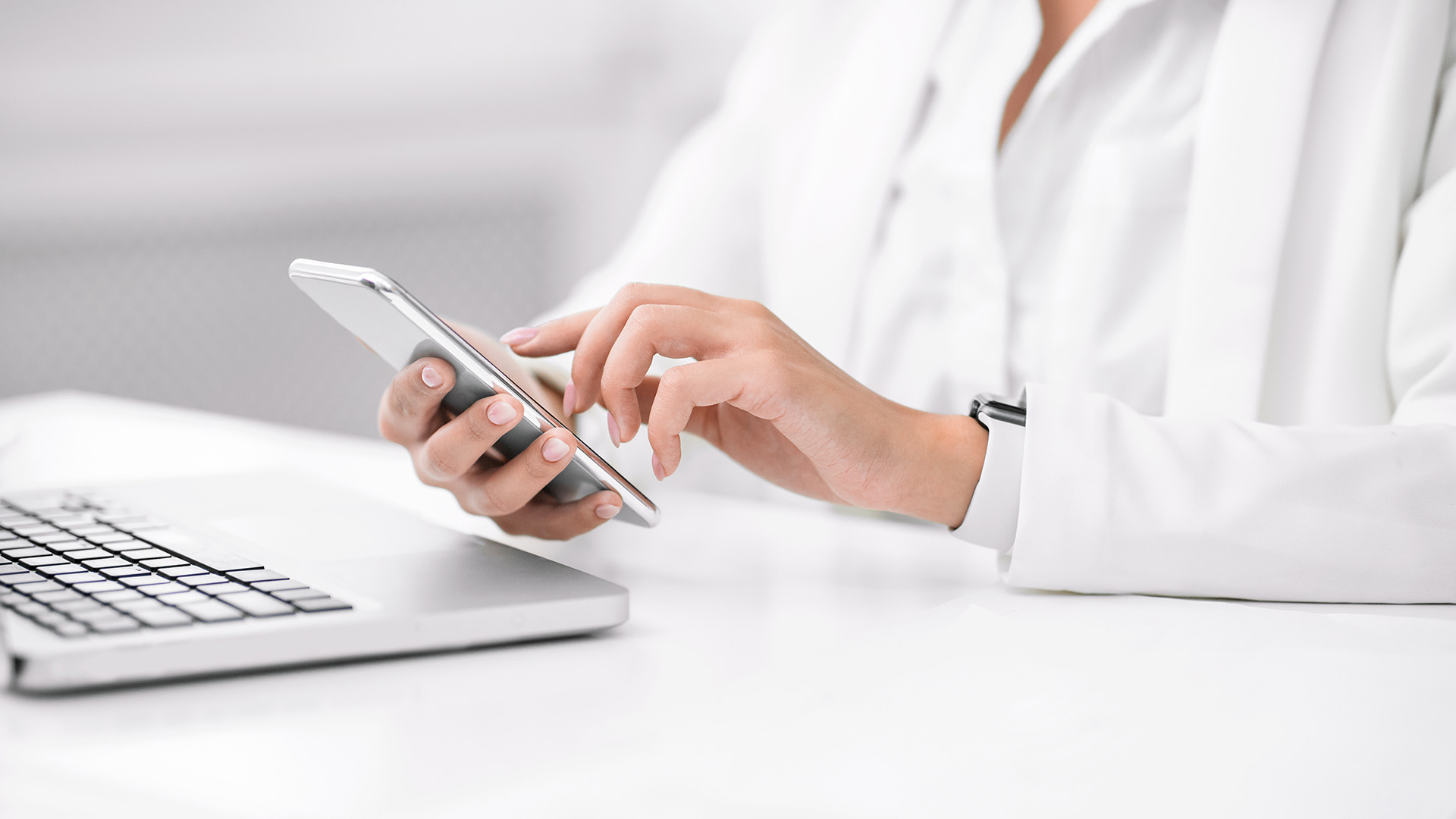 A person's hands using a smartphone while sitting at a desk with a laptop, set against a blurred background of an indoor office setting.