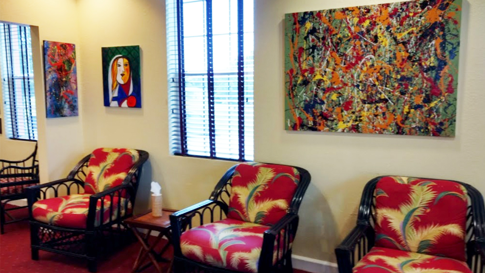 An art gallery interior displaying a variety of colorful abstract paintings on the wall, with empty chairs and a bench in front of them.