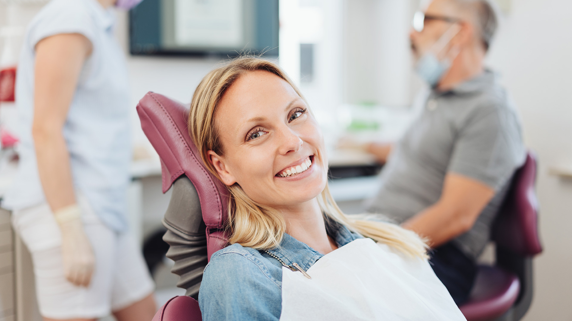 An adult woman with blonde hair is seated in a dental chair, smiling at the camera, while wearing an eye mask.