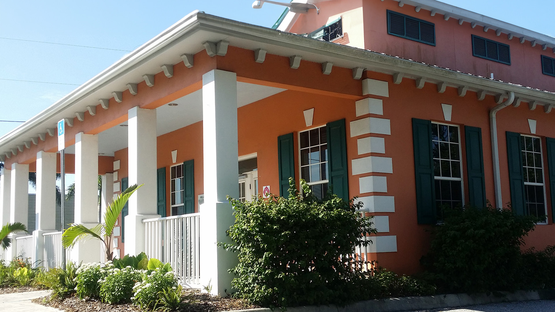 The image shows a two-story building with a peach exterior, a white roof, and a covered porch. There are columns supporting the porch roof, and the structure features a gabled roof with asphalt shingles. The front of the building has a prominent entrance with a set of double doors beneath an overhang supported by beams.