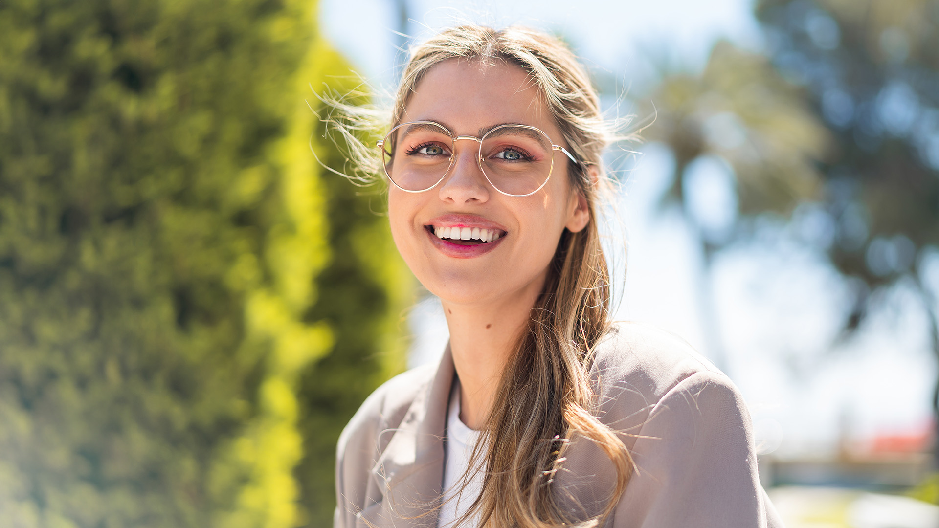 The image shows a woman with sunglasses, smiling at the camera, wearing a light-colored top and standing outdoors in front of a tree.