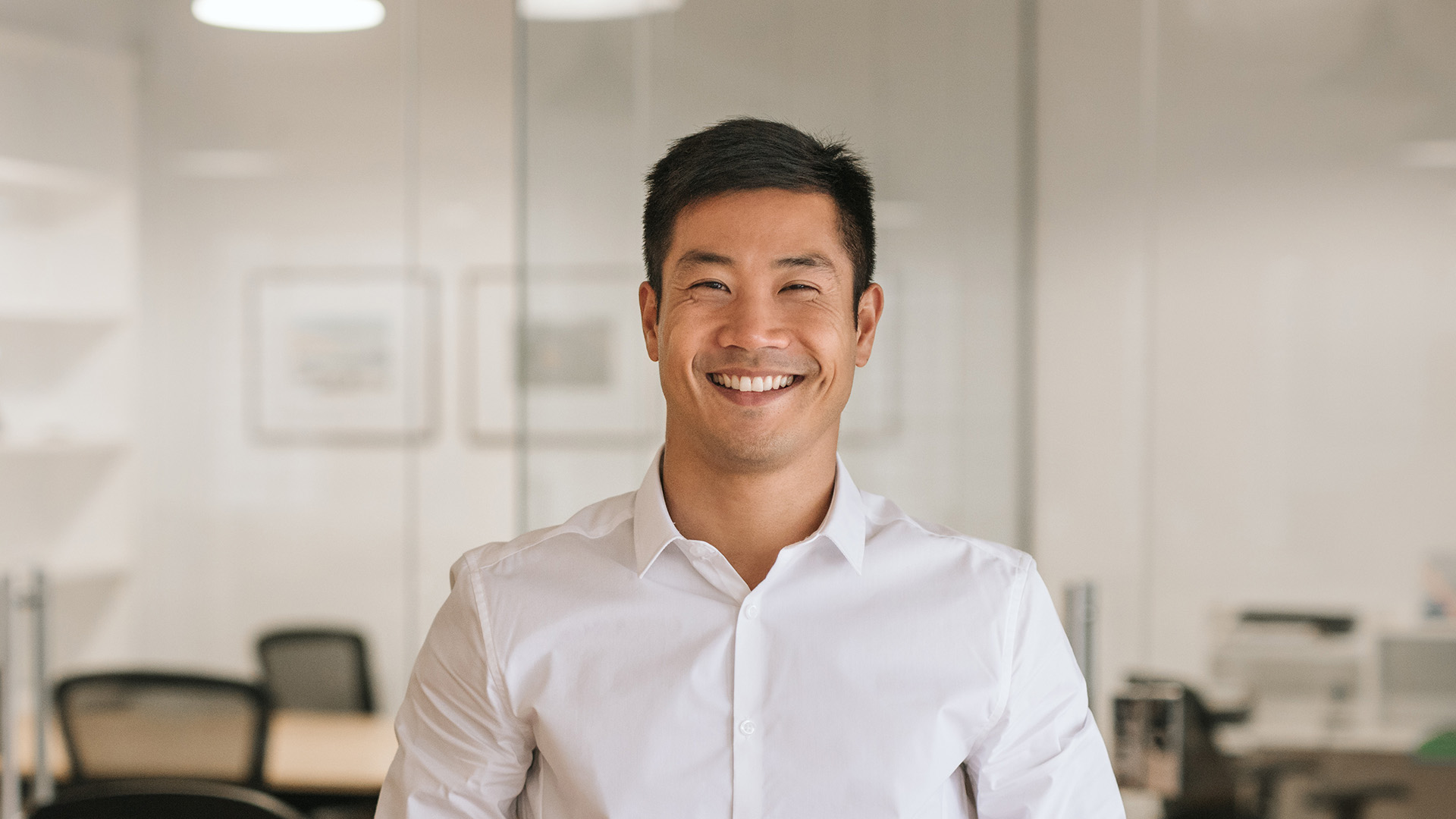 The image shows a man sitting at a desk in an office environment, smiling and looking directly at the camera.