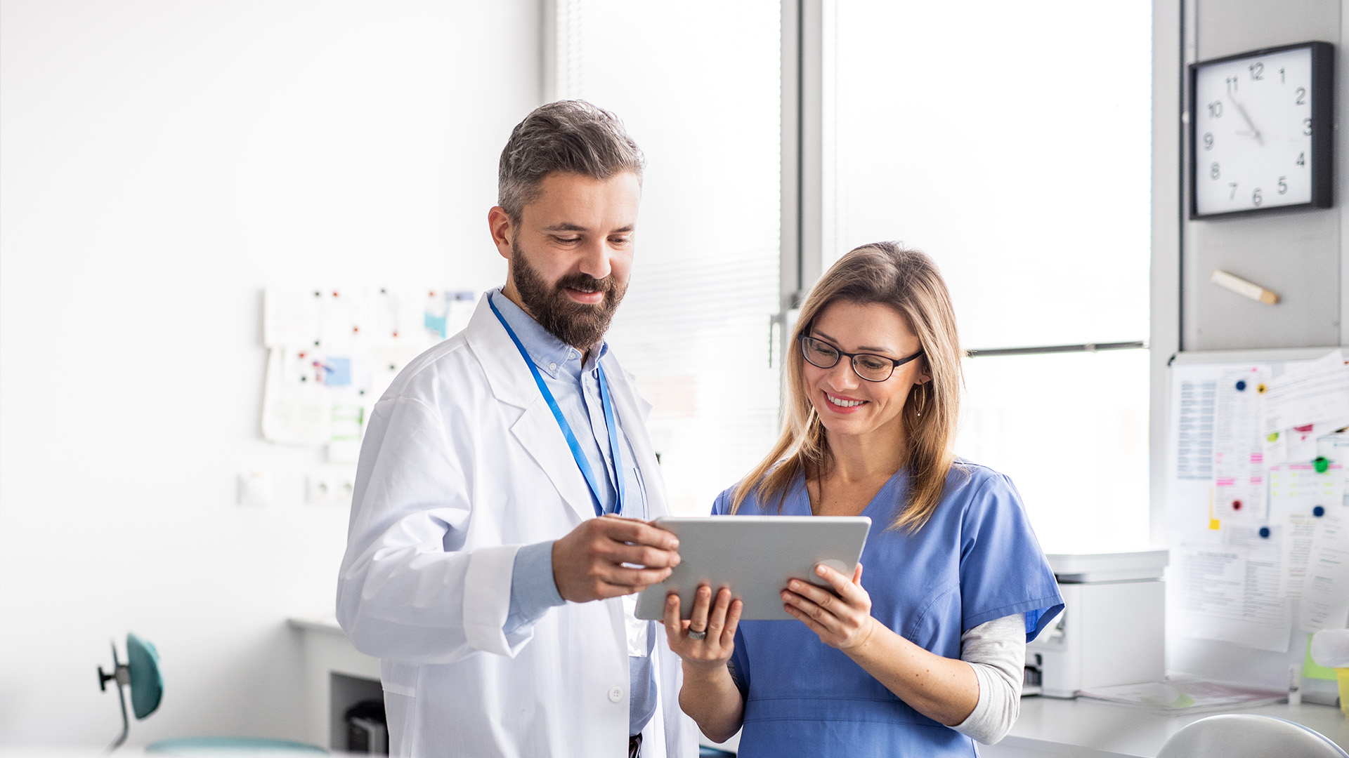 A man and a woman, both dressed in medical attire, are interacting with a tablet computer in an office setting.