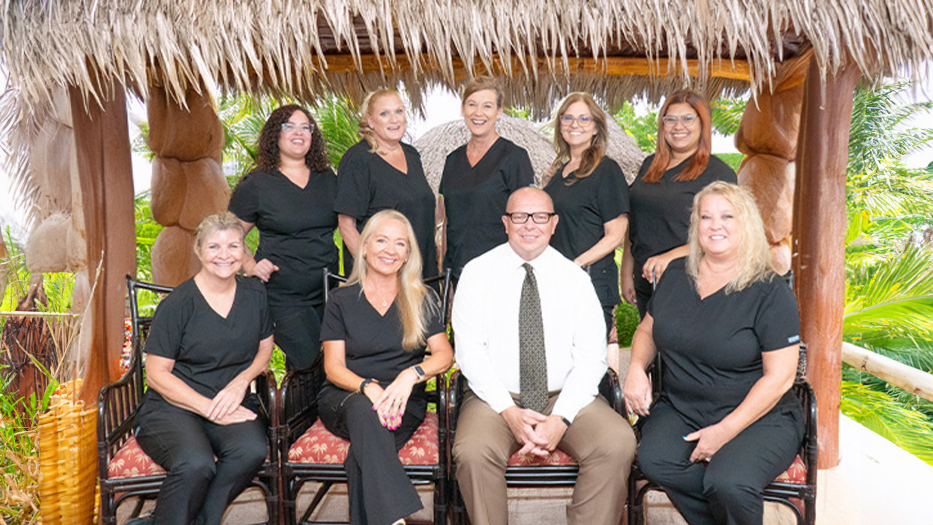A group of people, including a man standing in front, posing for a photo with a thatched roof structure behind them.