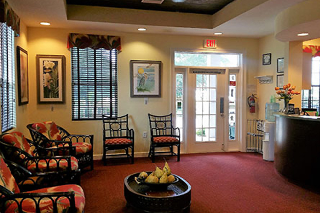 The image shows an interior view of a lobby or waiting area with a comfortable seating arrangement, including chairs and sofas, arranged around a central table. The room has a warm color scheme with beige walls and wood flooring, and there are framed pictures on the wall. A potted plant is visible in the corner, adding to the welcoming atmosphere.