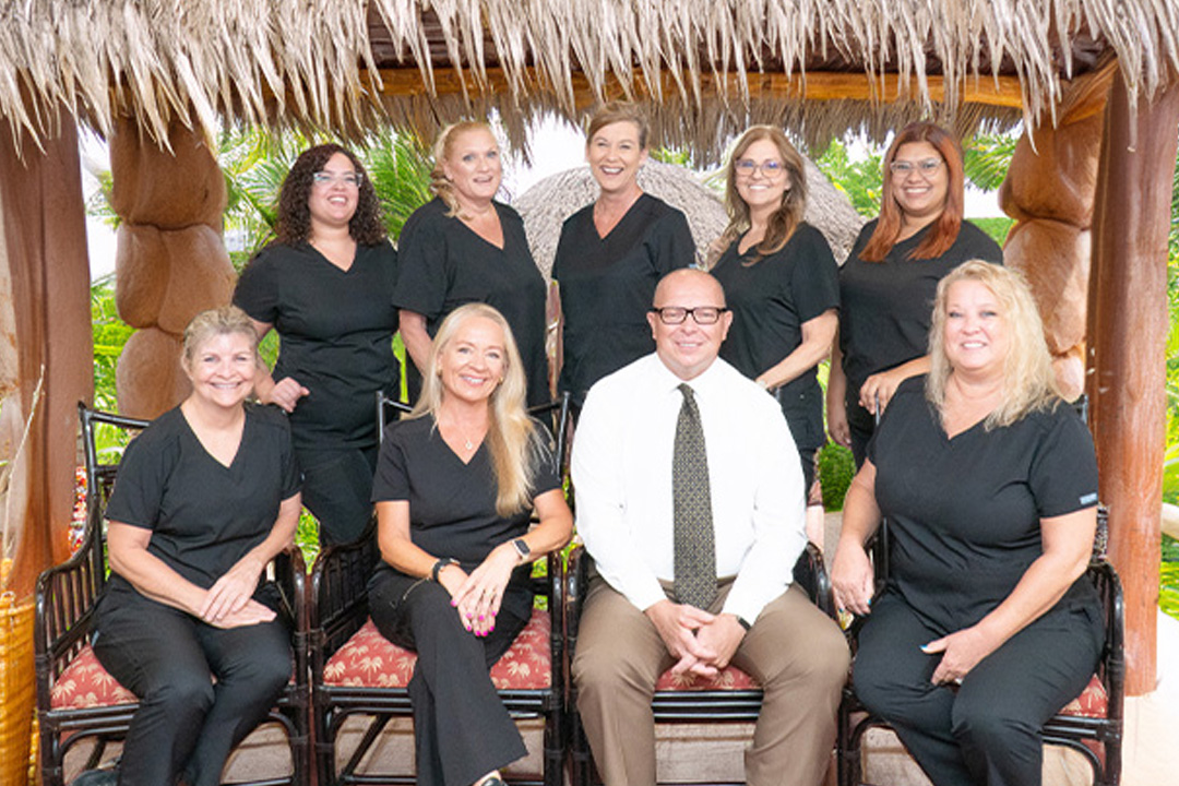 A group of seven women and one man in scrubs pose for a photo under a thatched roof hut.