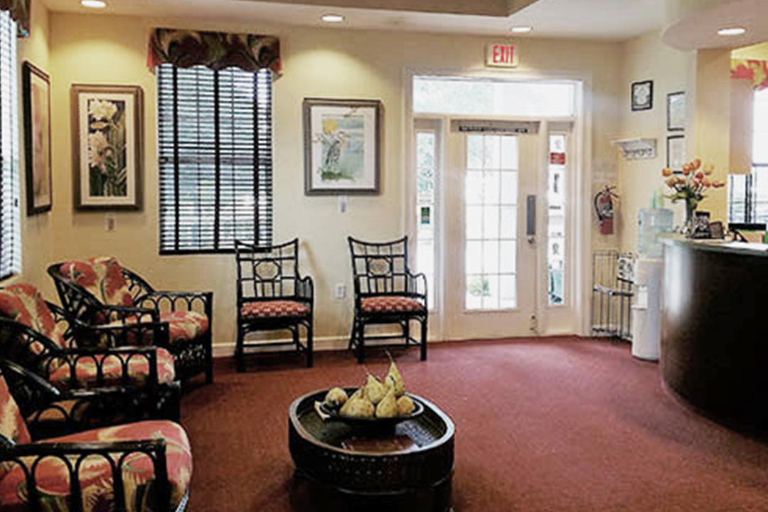 The image shows an interior view of a well-lit room with a waiting area, featuring comfortable seating arrangements, a reception desk, and framed art on the walls.