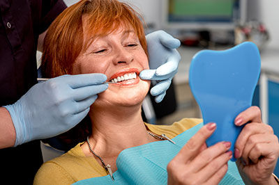 The image depicts a woman in a dental office, sitting in a chair with her mouth open, receiving dental care. She is holding a blue object that appears to be a dental appliance or model, and there are medical professionals present, one of whom is wearing gloves and adjusting the appliance. The setting includes dental equipment and a professional atmosphere.