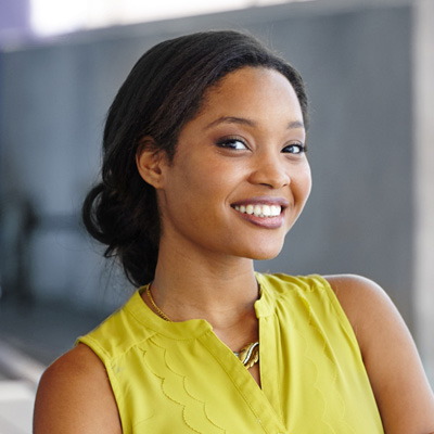 The image shows a woman with dark hair, wearing a yellow top and a necklace, smiling at the camera.