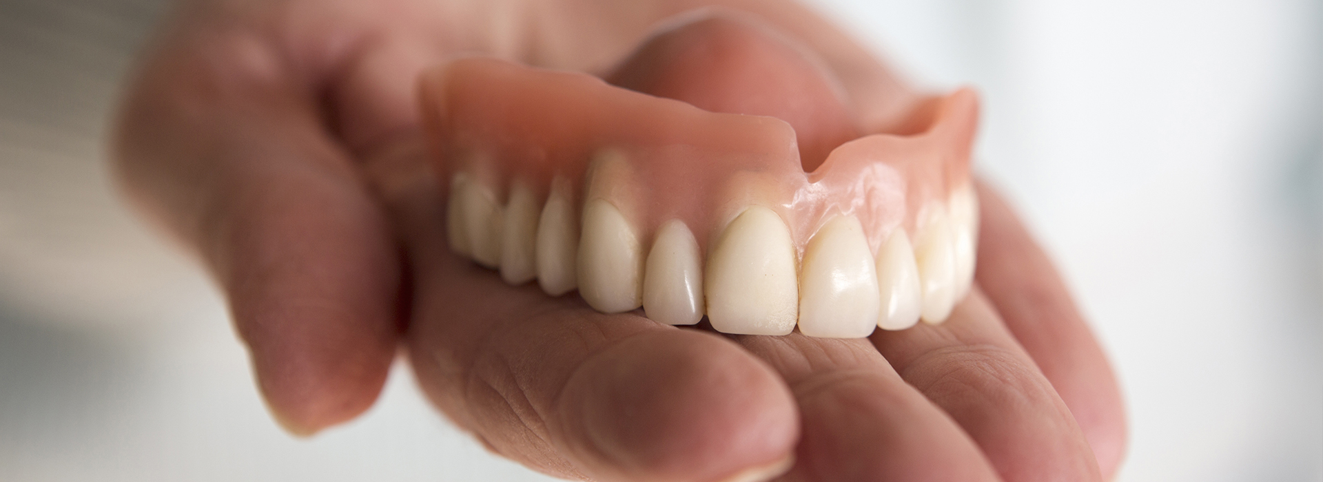 A person holding a set of dentures with their left hand, showcasing the prosthetic teeth against a neutral background.