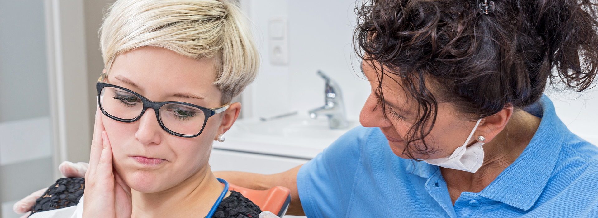 The image shows a woman with short hair, wearing glasses and a blue shirt, sitting in a dental chair with her eyes closed as she receives dental treatment. A female dentist is attending to her, holding a dental instrument near the patient's mouth.