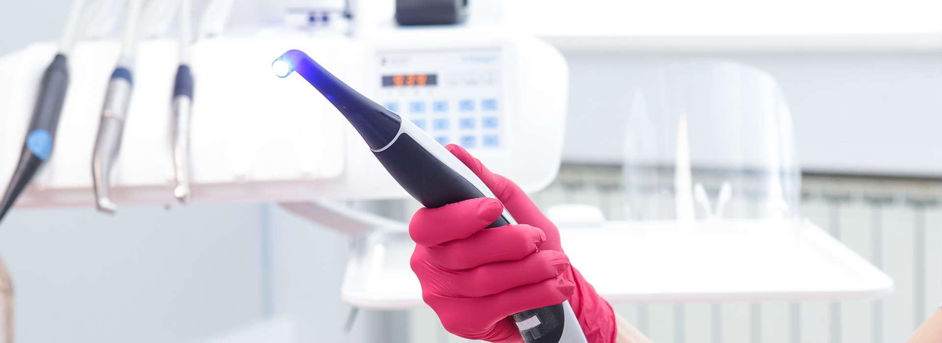The image shows a person wearing pink gloves and holding a blue device, possibly an electronic tool or gadget, in front of them. They appear to be in a professional setting with medical equipment in the background.