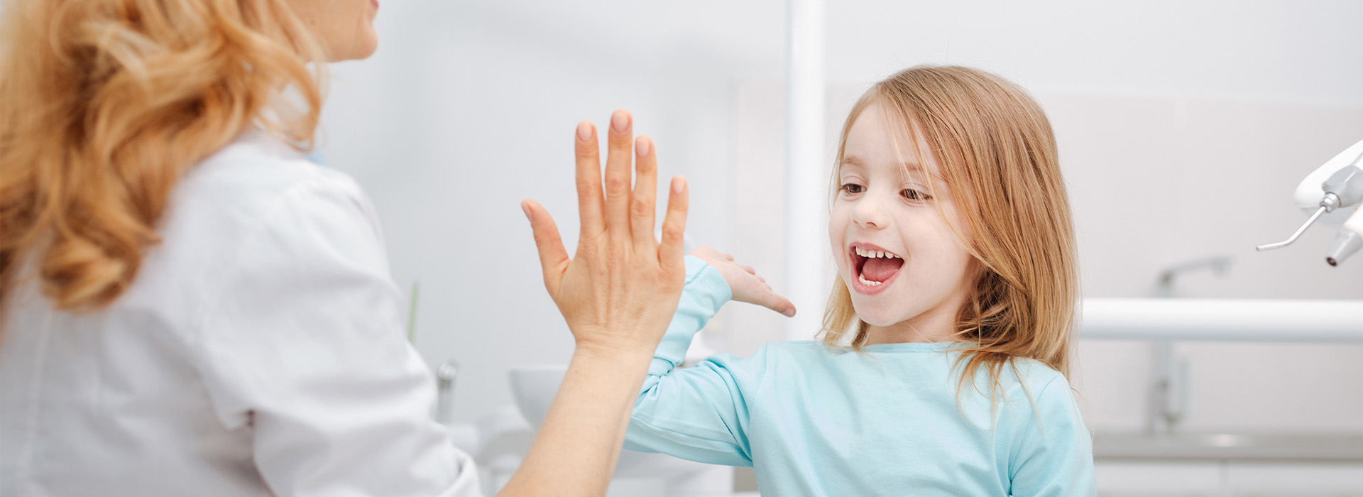A young girl and an adult woman in a dental office, the girl is seated in the dentist's chair with her hand raised, smiling at the camera while the woman stands beside her, also smiling, both are looking towards the camera.