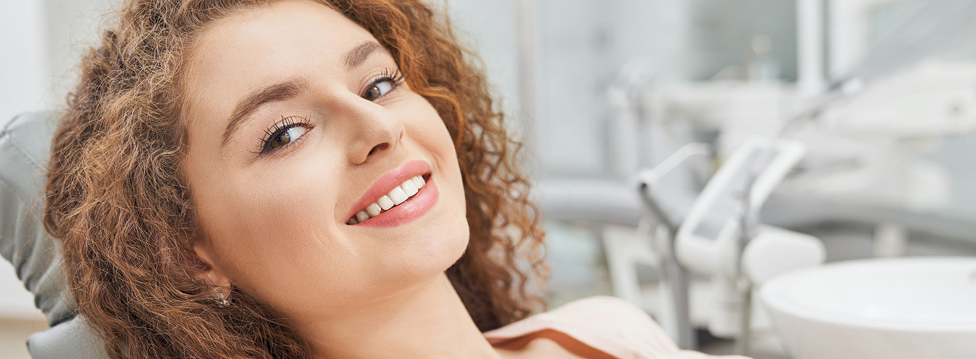 The image depicts a smiling woman with curly hair, sitting in a dental chair, looking at the camera.