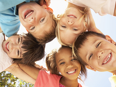 A group of six children, including three boys and three girls, smiling at the camera from a sunny outdoor setting.