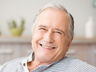 The image shows an elderly man with white hair, wearing a blue shirt and a black tie, smiling at the camera.
