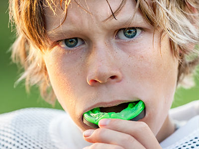 The image shows a young person with blonde hair and blue eyes, wearing a football uniform, holding a green object that appears to be an instrument or toy, possibly related to music, with their mouth open as if in the middle of playing it.